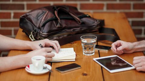 Zwei Menschen sitzen gemeinsam an einem Tisch mit Getränken, einer Ledertasche und Handys auf dem Tisch